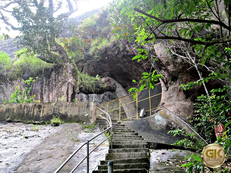 Concrete steps with steel railings climbing to a yellow grille under a large granite rock overhang, with trees and grass around it, at the Kuthirayala cave temple